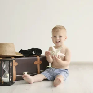 Petit garçon assis près d’une valise vintage avec un chapeau, des jumelles et un sablier dans une ambiance douce et minimaliste.