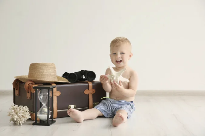 Petit garçon assis près d’une valise vintage avec un chapeau, des jumelles et un sablier dans une ambiance douce et minimaliste.