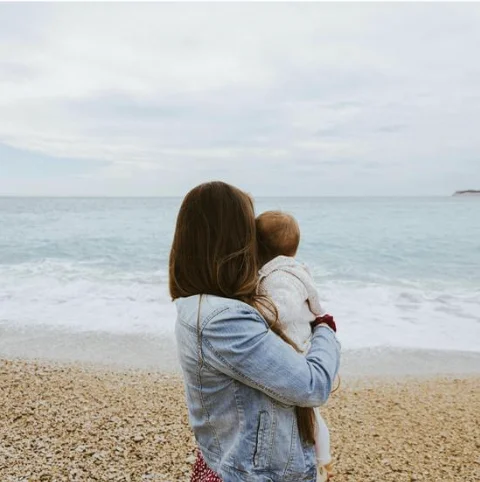 Femme de dos en veste en jean tenant un jeune enfant dans ses bras face à la mer sur une plage de galets par temps nuageux
